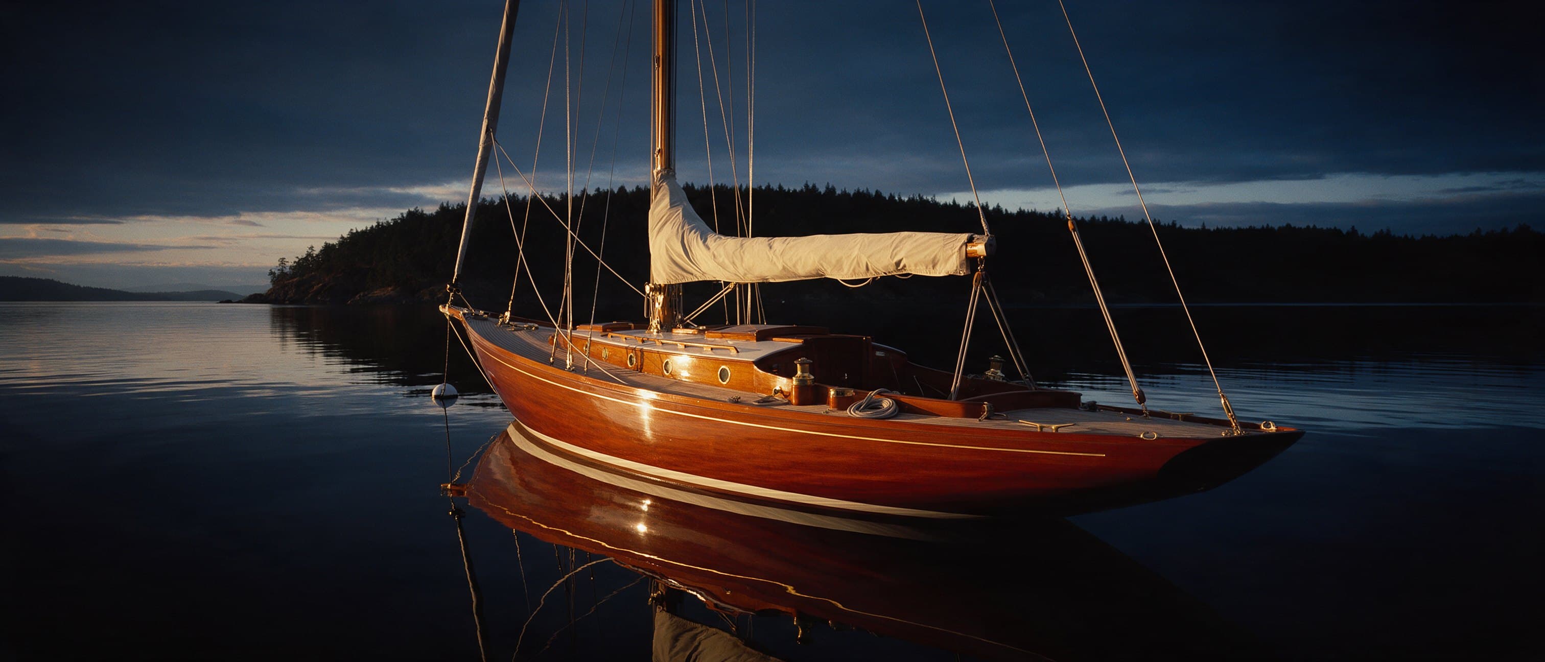 A varnished wooden sloop lying at a private mooring at dusk, its deck empty and its reflection unbroken on still water.